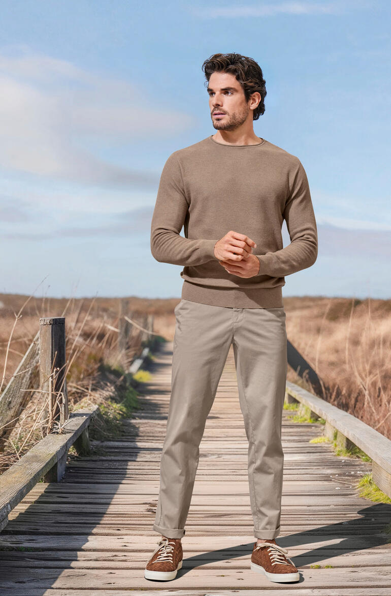 Taupe Mann in beigem Pullover und hellen Hosen auf einem Holzweg