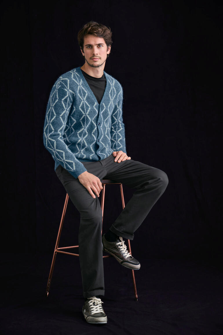 Schwarz Mann sitzt auf Barhocker in blauem Brushed-Alpaka-Cardigan mit Rautenmuster.