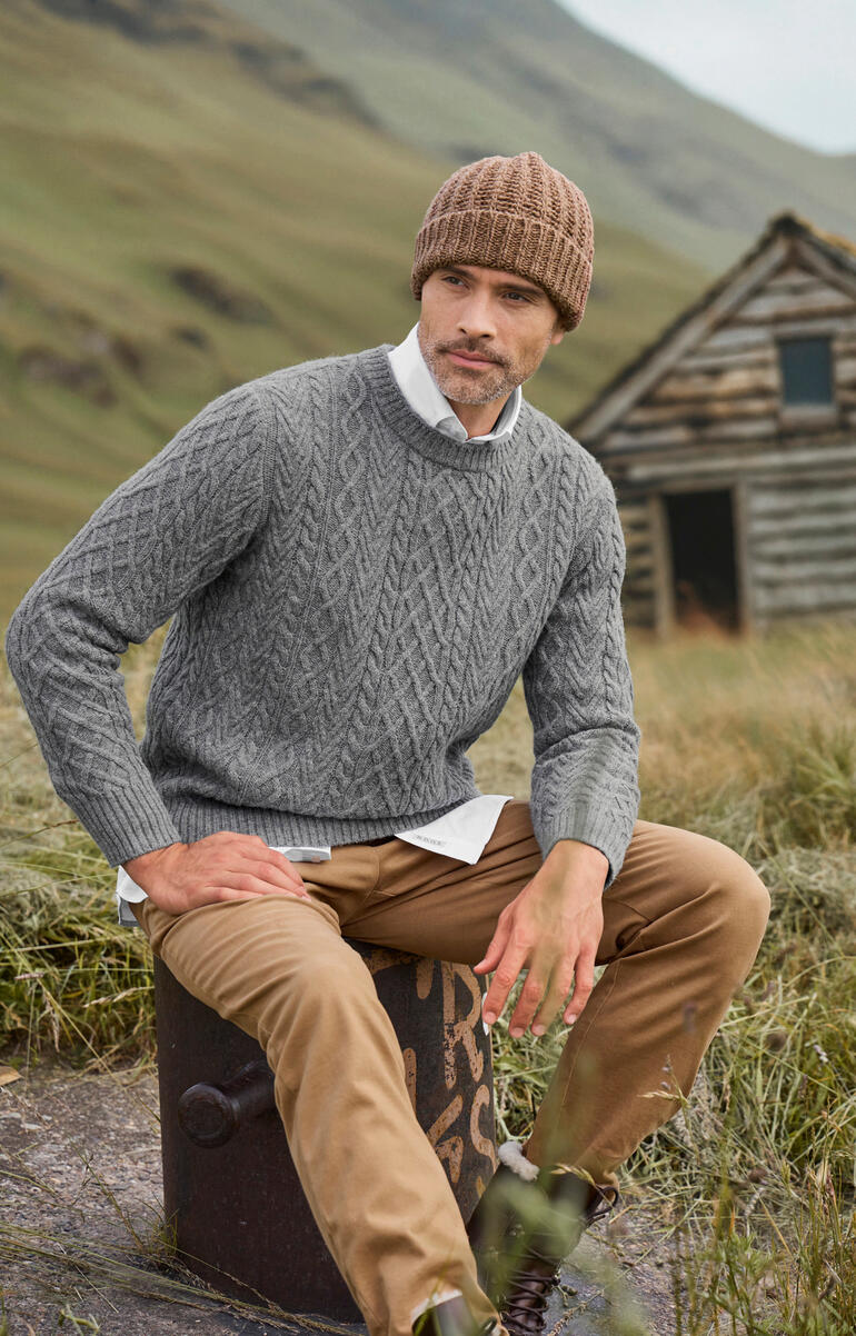 Duneen Ireland Aran-Pullover Mann sitzt auf einem Stein, trägt einen grauen Aran-Pullover und Mütze.