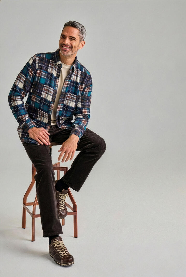 Mann sitzt auf einem Hocker, trägt ein kariertes Flanellhemd in Blau und Braun.