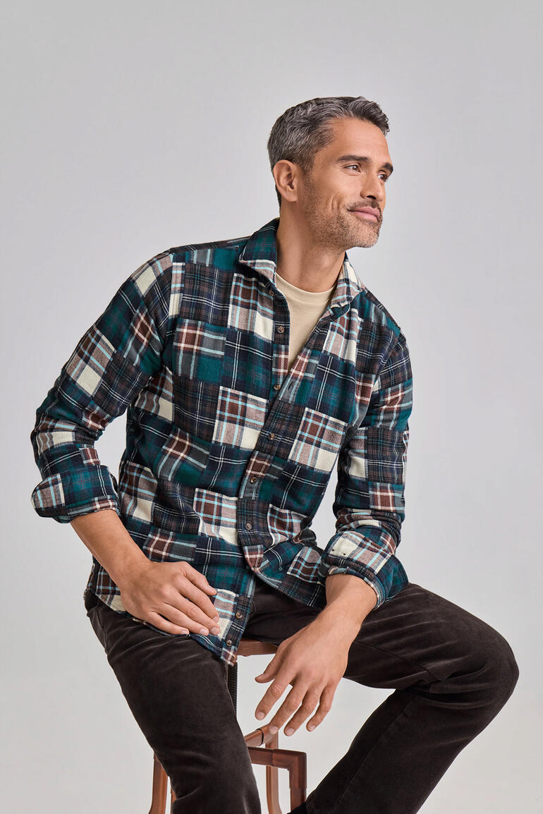 DU4 Patchwork-Flanellhemd Mann trägt ein DU4 Patchwork-Flanellhemd mit Karomuster, sitzt auf einem Hocker.