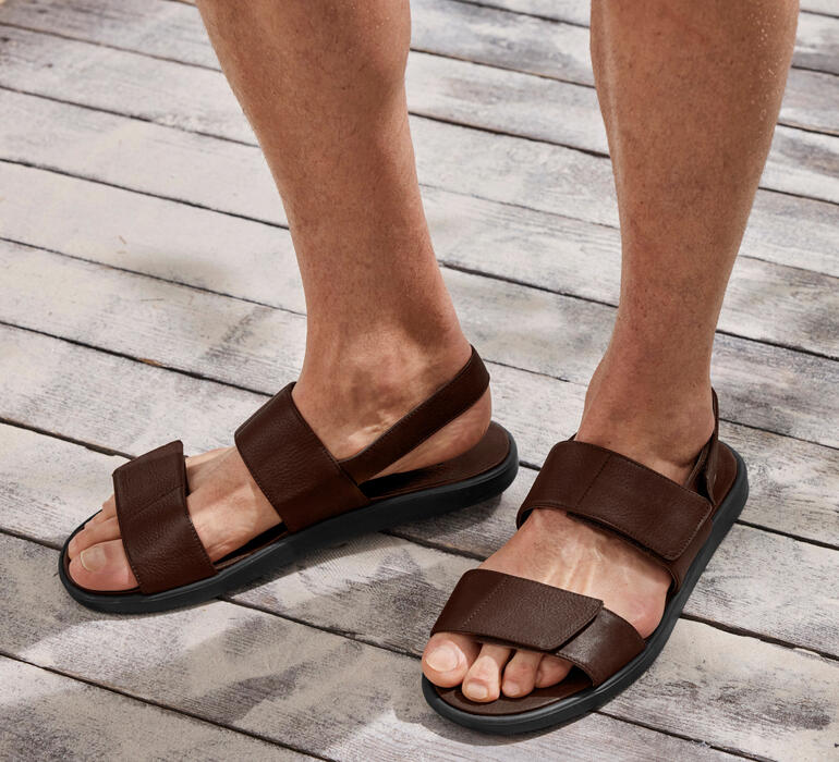 Braunes Leder-Sandale mit zwei Riemen an einem Fuß auf einem Holzfußboden.