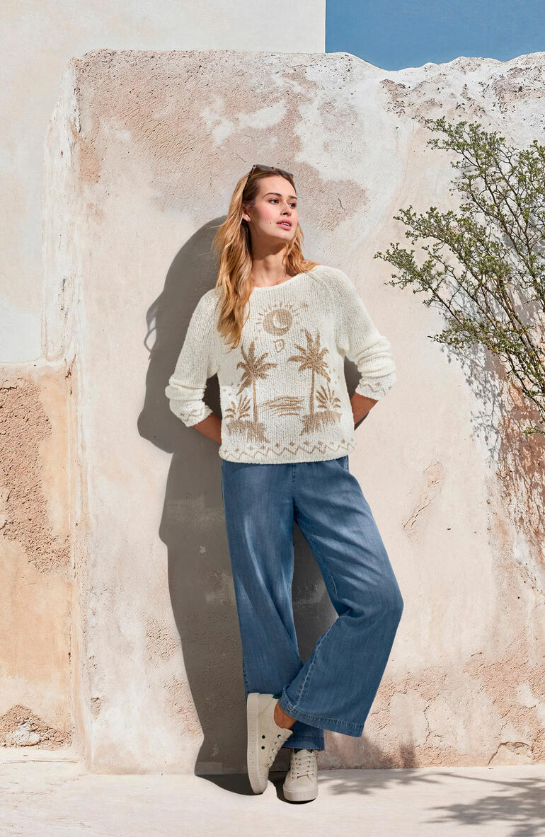 Frau in cremefarbenem Pullover und hellblauen Wide-leg-Jeans, vor einer Wand.