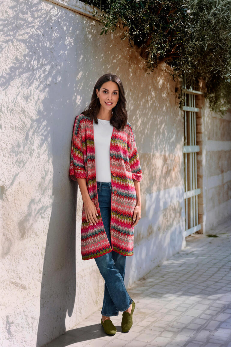IVKO Jacquard-Langcardigan Frau in einem bunten, gestrickten Langcardigan mit farbigen Rautenmustern, steht an einer Wand.