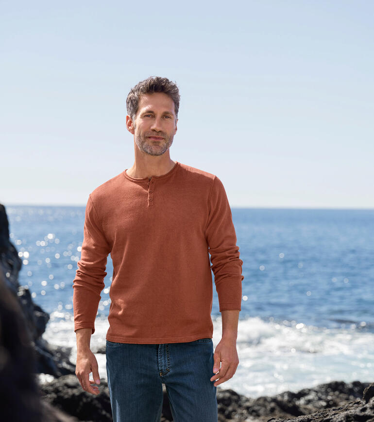 Ein Mann steht am Strand in einem rostroten Ragman Pullover und Jeans.