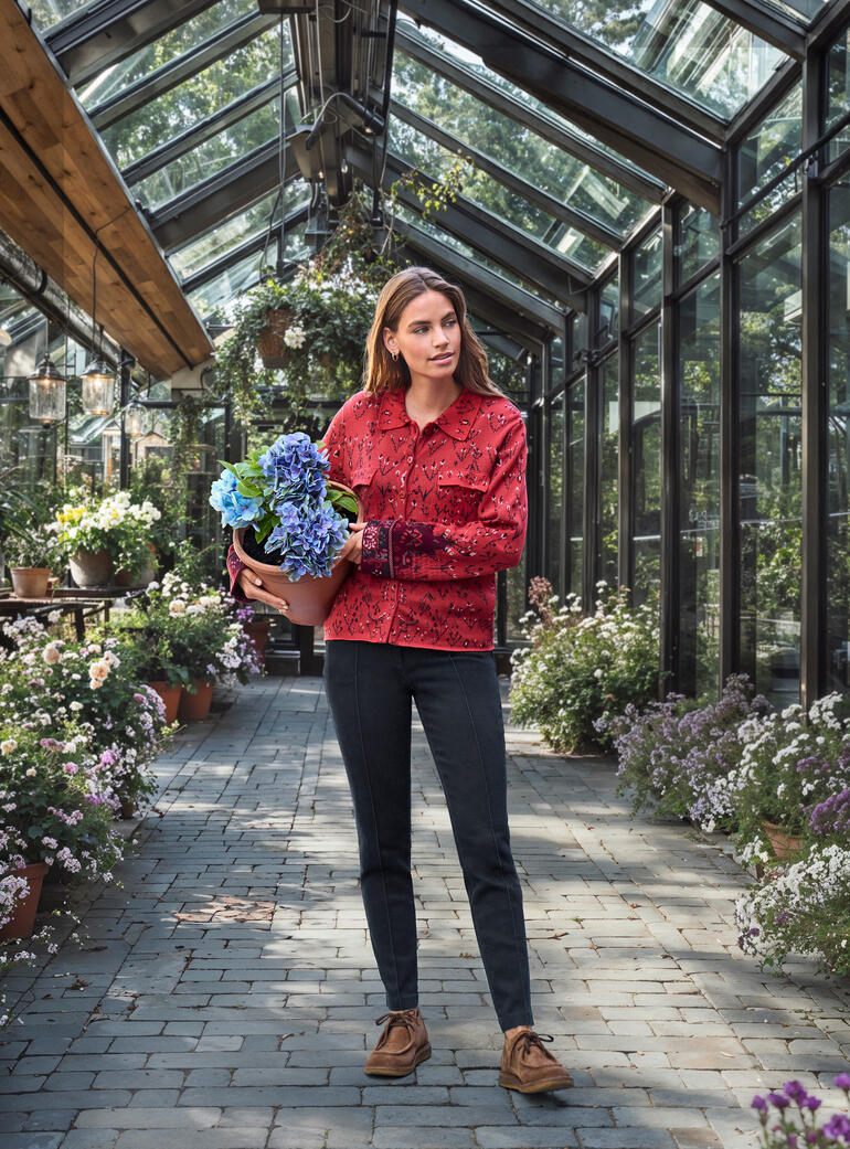 Frau in blumenmusternder Jacquard-Strickbluse in einem Gewächshaus mit Pflanzen.