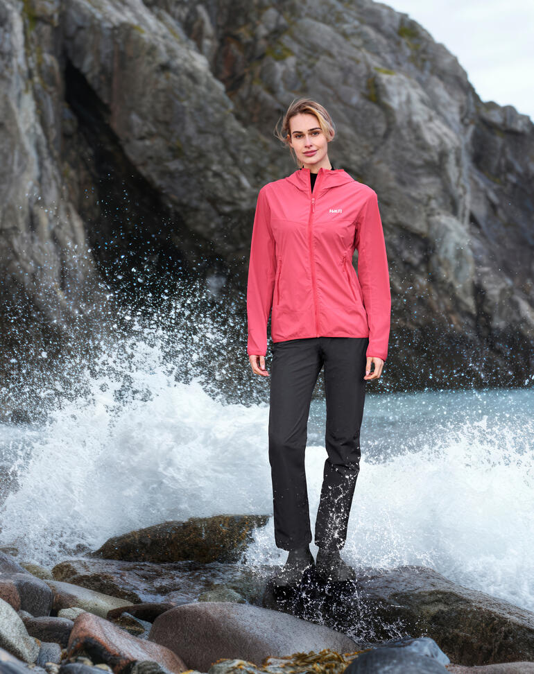 Frau in pinker Softshelljacke und schwarzer Hose, steht am Strand mit Wellen im Hintergrund.