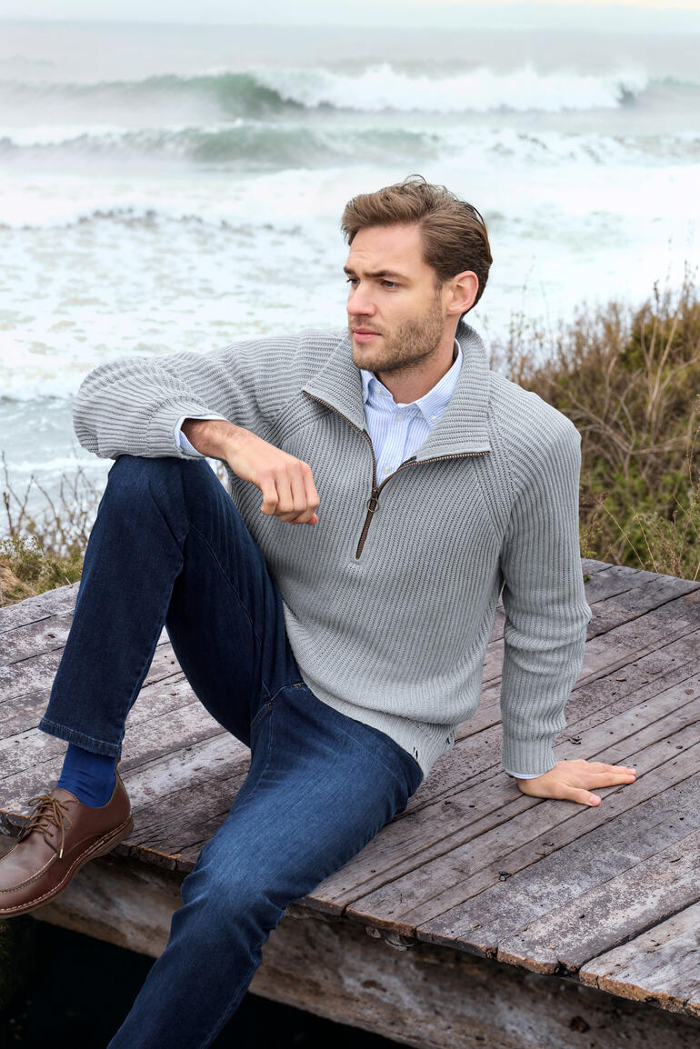 Mann im Rippstrick-Troyer sitzt am Strand und blickt auf das Meer.