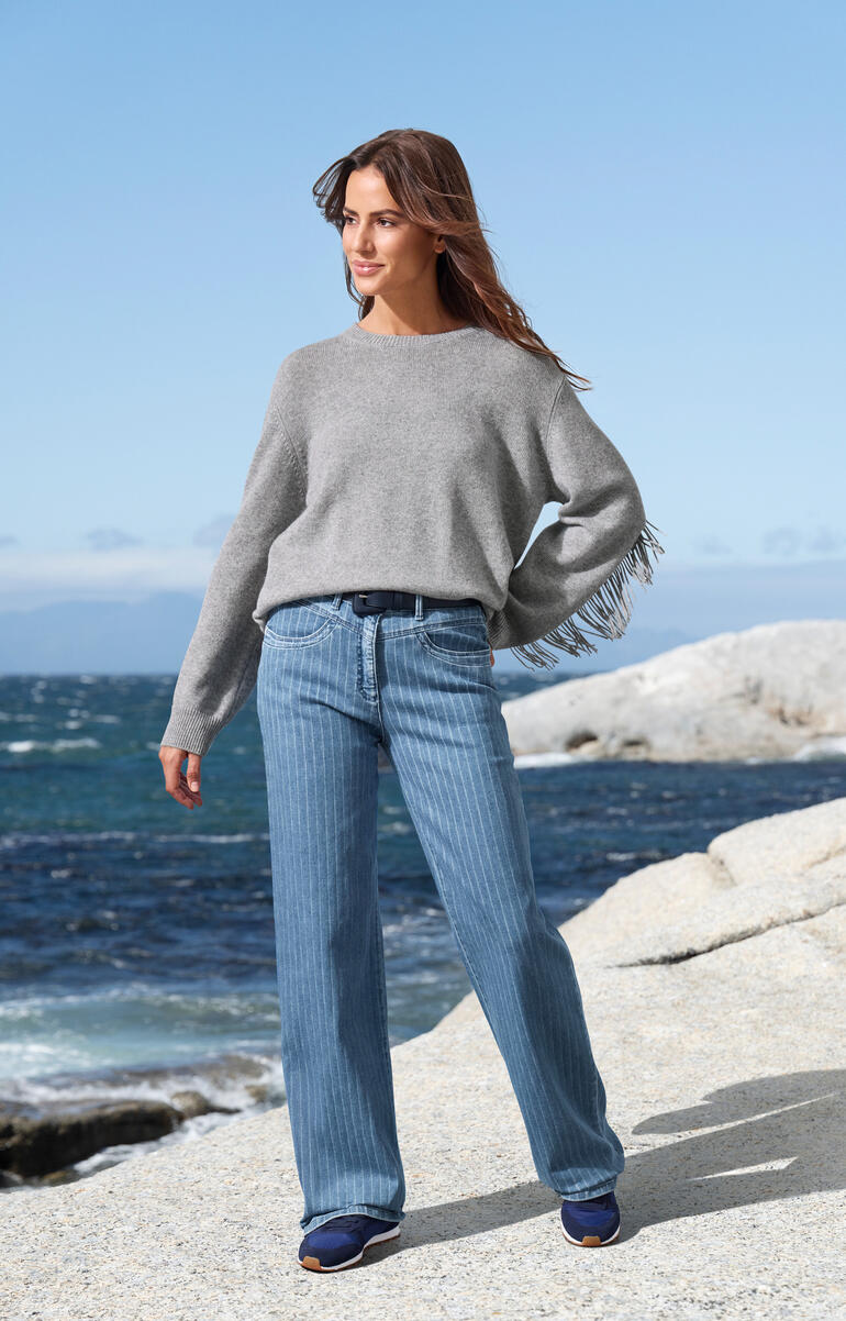 Grauer Pulli mit Fransen, lässig über Jeans getragen am Strand.