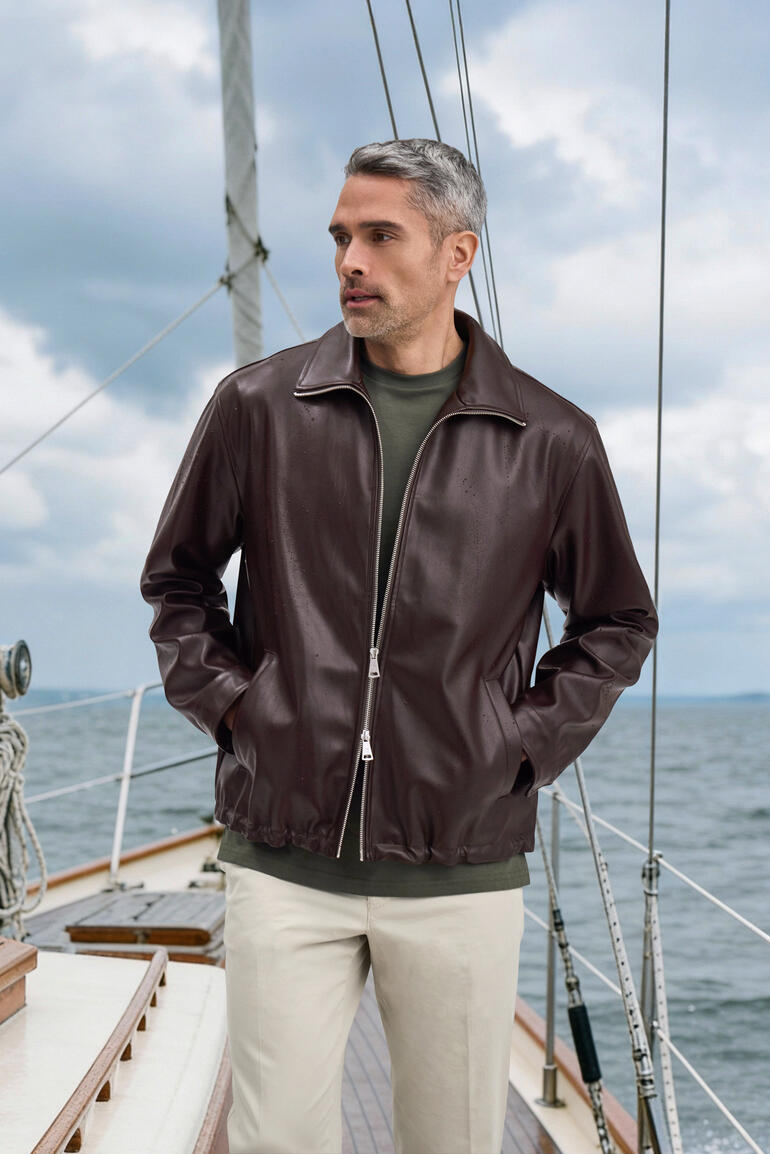Mann in brauner Kunstlederjacke mit grauem Pullover auf dem Deck eines Segelboots
