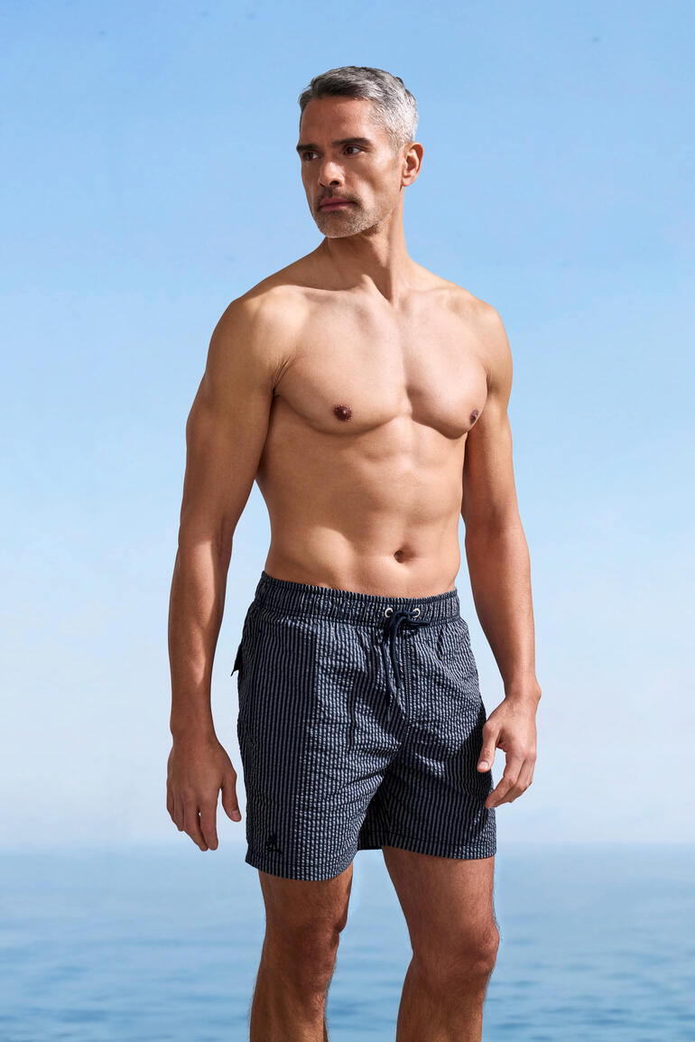 Mann trägt Streifenbadeshorts am Strand, mit freiem Oberkörper.