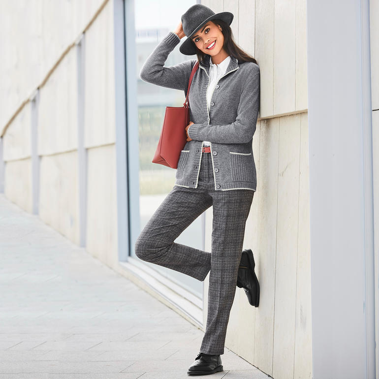 Grau Frau in grauem Kaschmir-Cardigan mit schwarzem Hut und roter Tasche, lehnt an Wand.