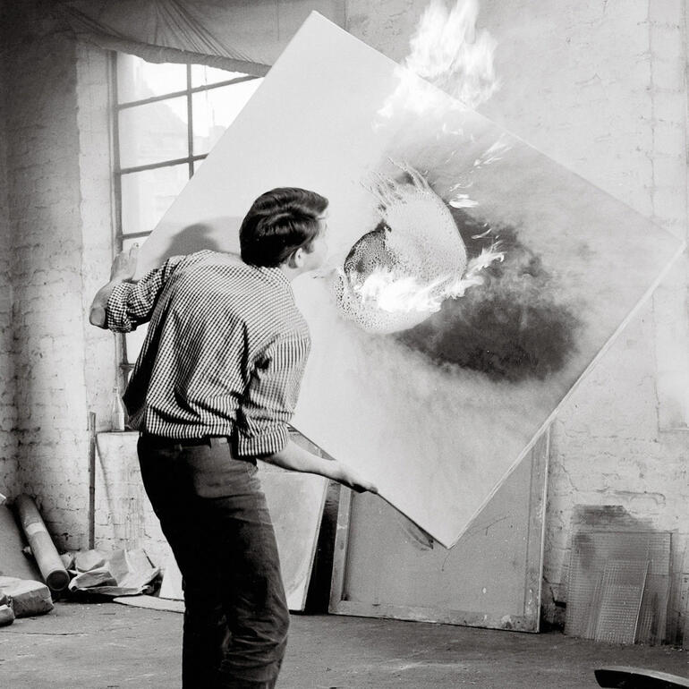 Durch Flammen gibt Otto Piene den Materialien der Bildfläche eine völlig veränderte Erscheinungsform. Künstler, der ein Kunstwerk mit Feuer hält, während Flammen auf der Leinwand zu sehen sind.