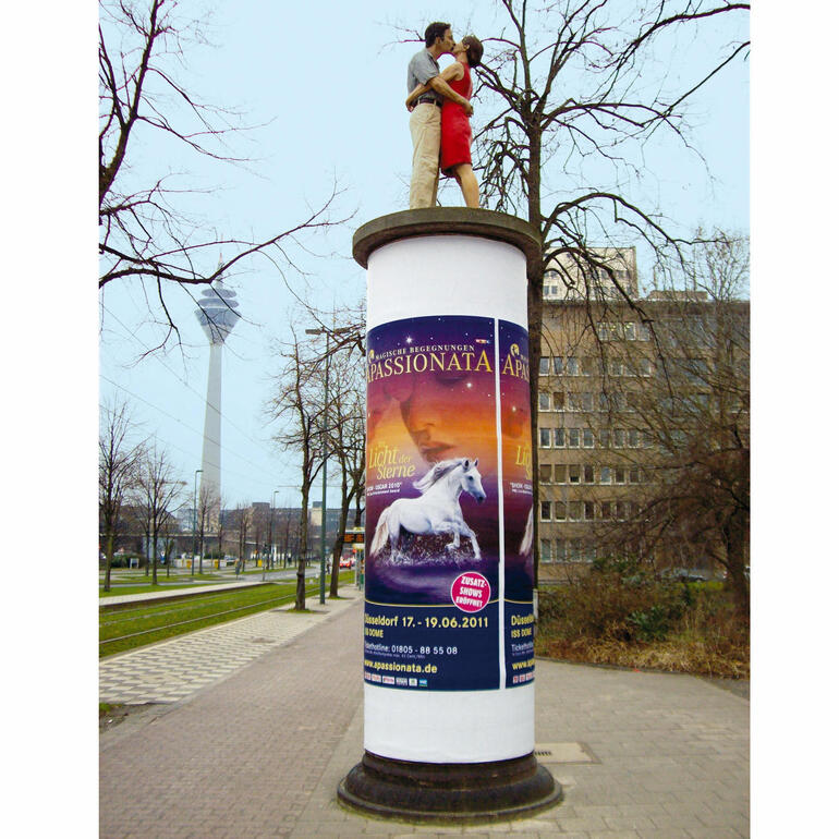 Die 1,90 m große Originalskulptur auf einer Litfass-Säule ist in der Düsseldorfer Innenstadt (Ecke Haroldstraße/Poststraße) zu sehen. Paar, das sich auf einer Säule in der Stadt umarmt, umgeben von Werbeplakaten.