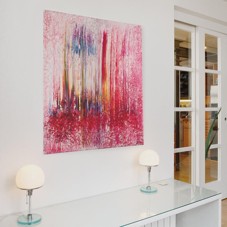 Aufgrund seiner farbigen Vielschichtigkeit entwickelt das Werk eine Eigendynamik, die jeden Raum bereichert. Abstraktes Wandbild in Pink- und Rottönen, hängt über einem modernen Tisch mit Lampen.