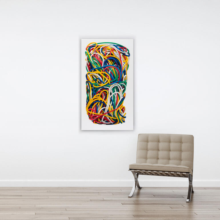 Die Gummibänder sind so realistisch gemalt, dass man erst auf den zweiten Blick erkennt, dass es keine Fotografie ist. Buntes Gummiband-Kunstwerk hängt an einer weißen Wand über einem Stuhl.