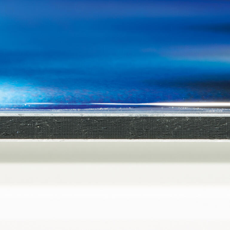 Das Werk auf DIBOND® wird mit einem klaren Kompositharz ummantelt, das die optische Tiefenwirkung verstärkt und die Farben intensiv leuchten lässt. Nahaufnahme der Kante eines Bildes mit glattem Finish.