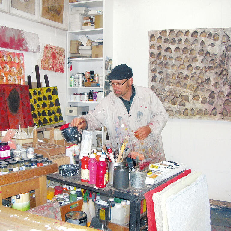 André Schweers in seinem Atelier. Künstler in seinem Atelier, umgeben von Farbtöpfen und Kunstwerken.