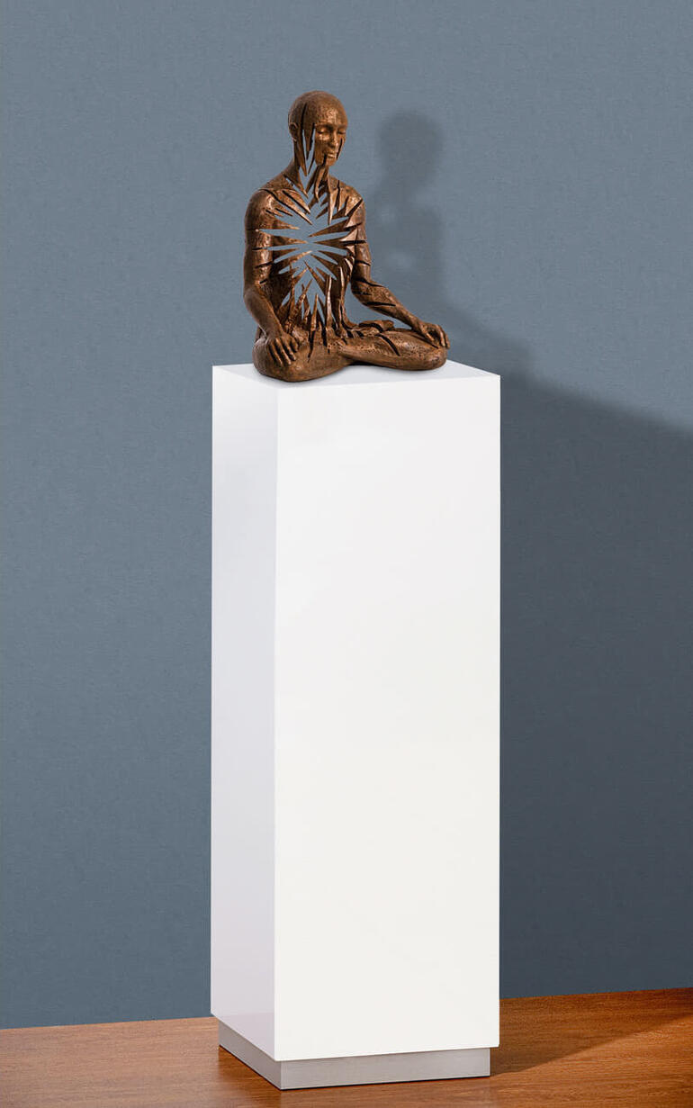 Weiße Säule mit bronzeartiger Meditationsskulptur auf einer Holzoberfläche.