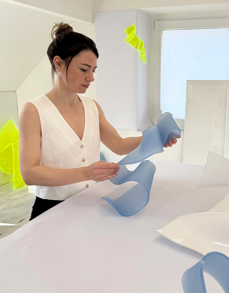 Frau mit kurzen Haaren formt farbige, transparente Kunststoffstreifen auf einem Tisch.