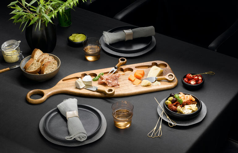 Design-Servierbrett aus Akazienholz mit Käse, Obst und Snacks auf einem dunklen Tisch.