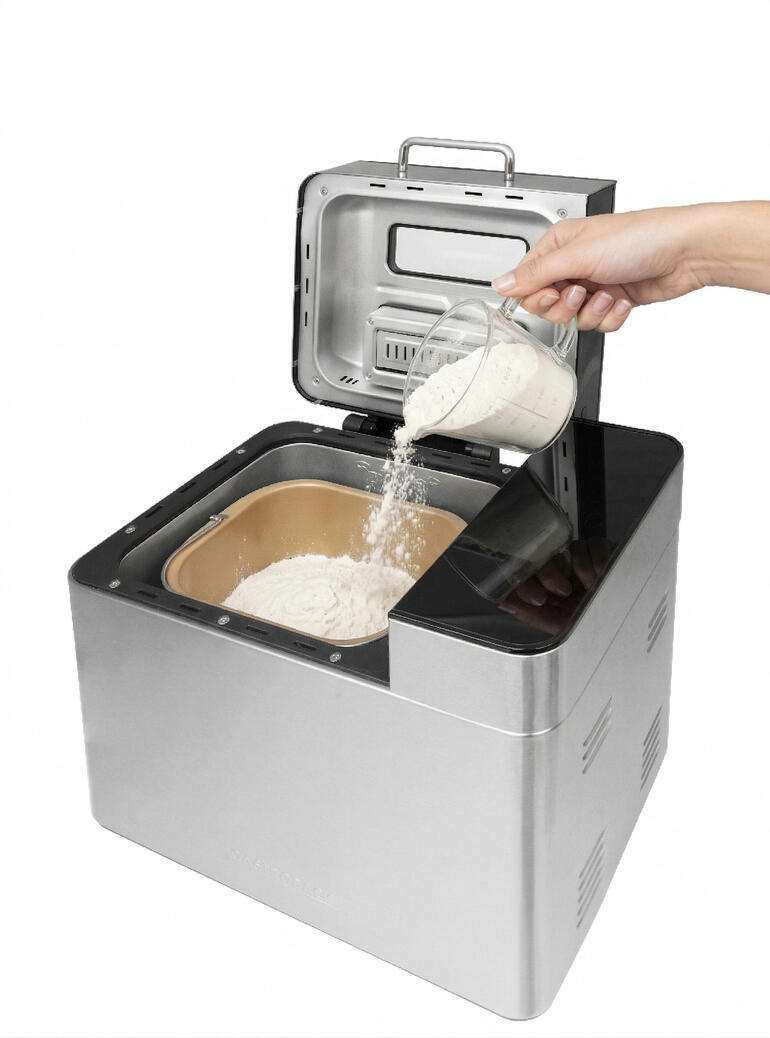 Hand, die Mehl in den geöffneten Brotbackautomaten schüttet.