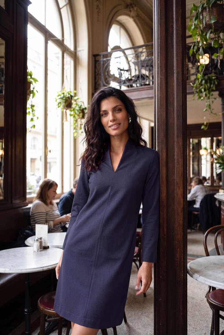 Frau in kleinem, blauen Kleid mit Kelchkragen in einem Café.