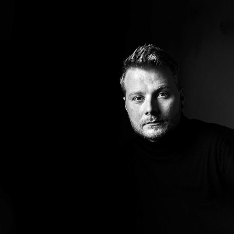 David Tollmann Mann im schwarzen Rollkragenpullover vor dunklem Hintergrund.